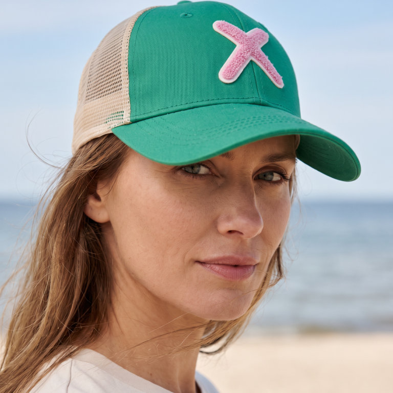 gen AI, eine Dame mit einem Unio Hamburg Basecap am Strand, Portraitaufnahme, Basecap grün, beige mit einem Fliederfarbenen Teddyfell X, bildformat 1:1